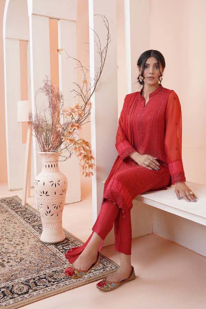 A woman in traditional red clothing sitting elegantly indoors, showcasing Pakistani fashion.
